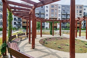 Una estructura de pérgola de madera con bancos, rodeada de un jardín ajardinado y edificios residenciales al fondo.