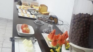 A buffet table with various food items including fruits, a cake, and a beverage dispenser. - Ami Hotel (Varzea Grande)