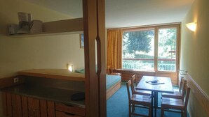 Ein Zimmer mit Etagenbett, einem Esstisch mit Stühlen, einem Fenster mit Vorhängen und einer kleinen Küchenzeile inklusive Kaffeemaschine.