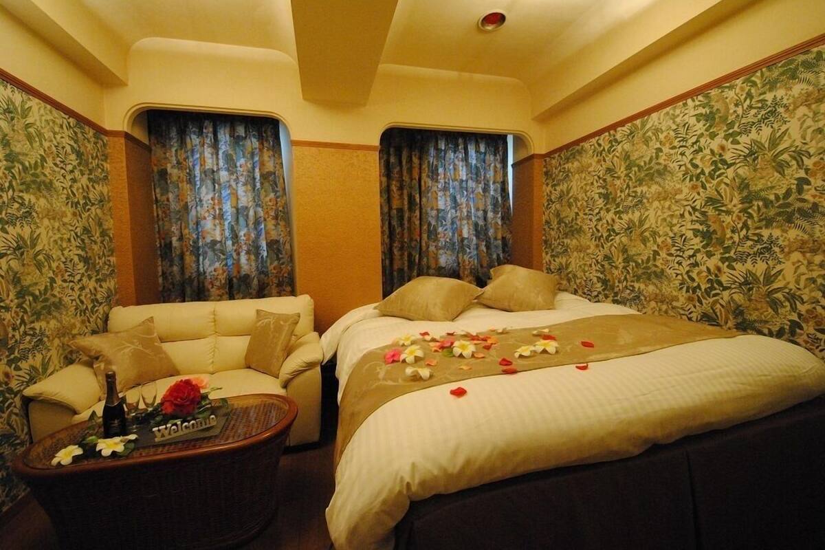 Una habitación de hotel con un sofá, una cama con colcha estampada de flores, una mesa de centro con una botella y flores, y cortinas con un estampado tropical.