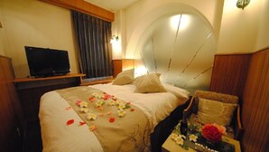 A hotel room with a bed, a television on a stand, a round headboard, and a small table with a welcome sign. - Hotel Marion (Suzuka)