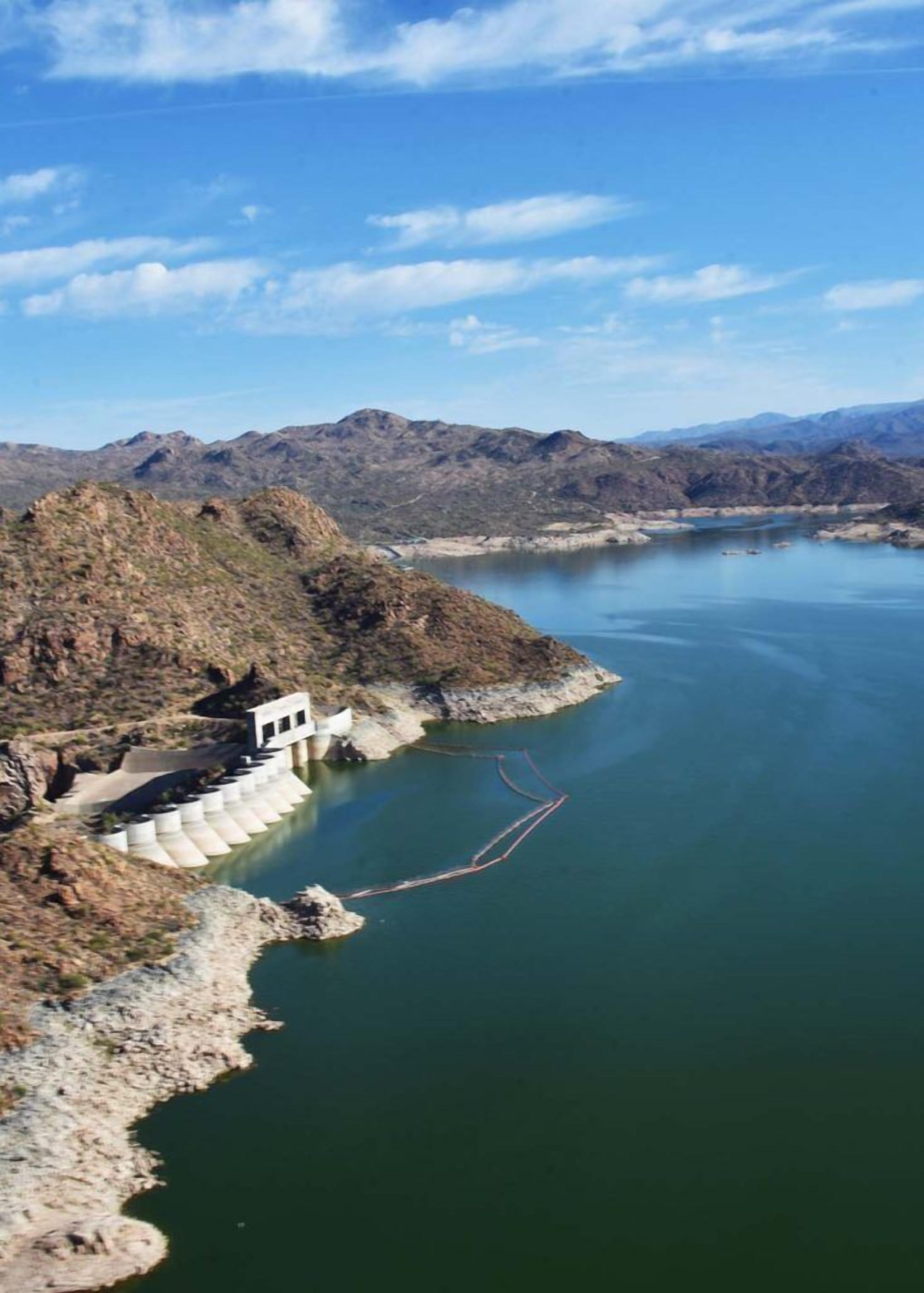 A dam with a small building on a hillside, a calm river, and distant mountains.