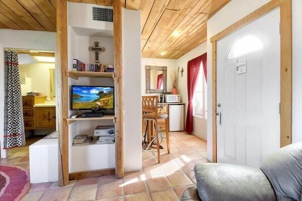 Un salon chaleureux avec un plafond en bois, un téléviseur, une bibliothèque, un coin repas avec une chaise et une table, et une porte blanche.