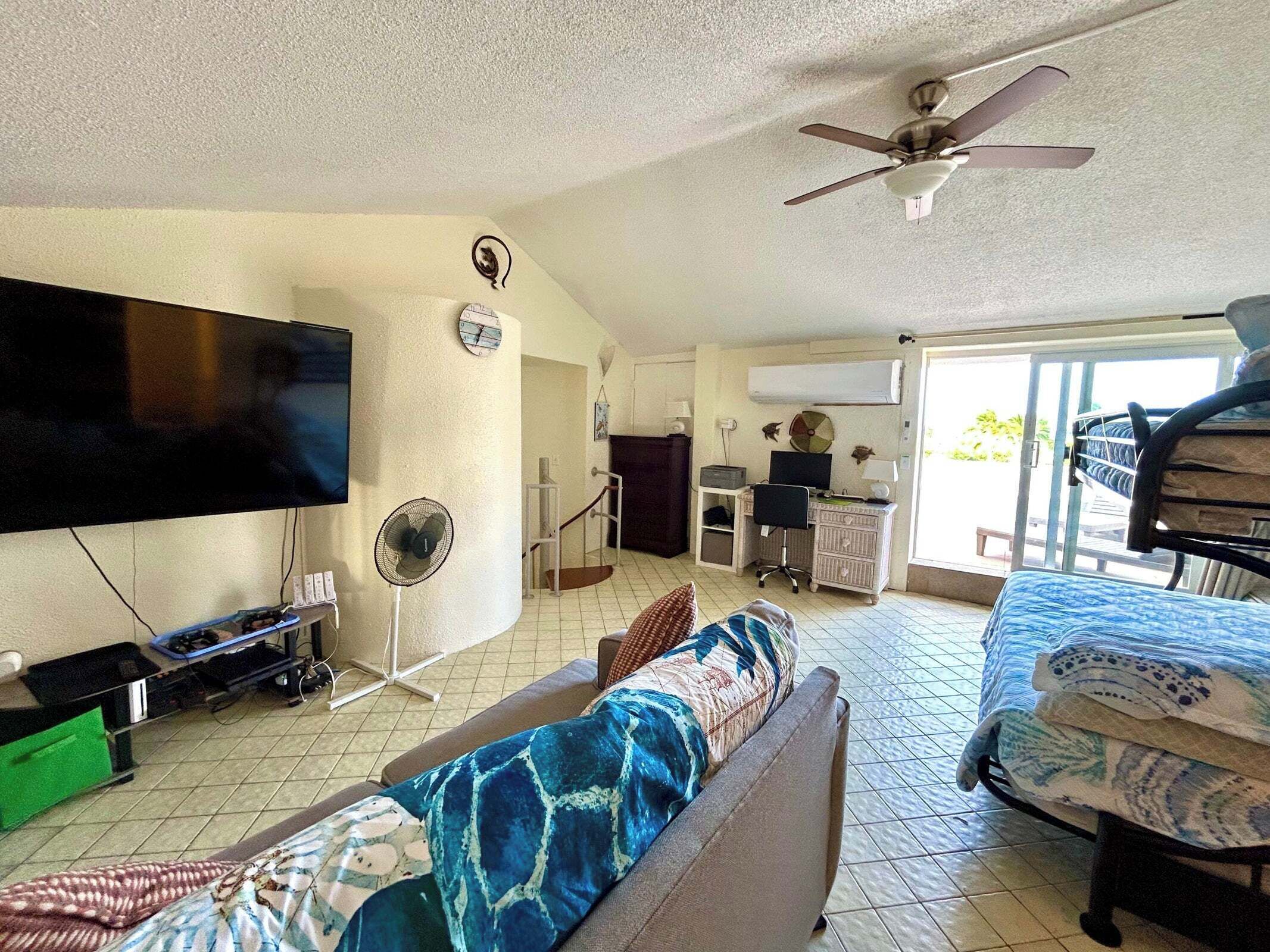 A spacious living room with a flat-screen TV, ceiling fan, and a bed with a patterned bedspread.