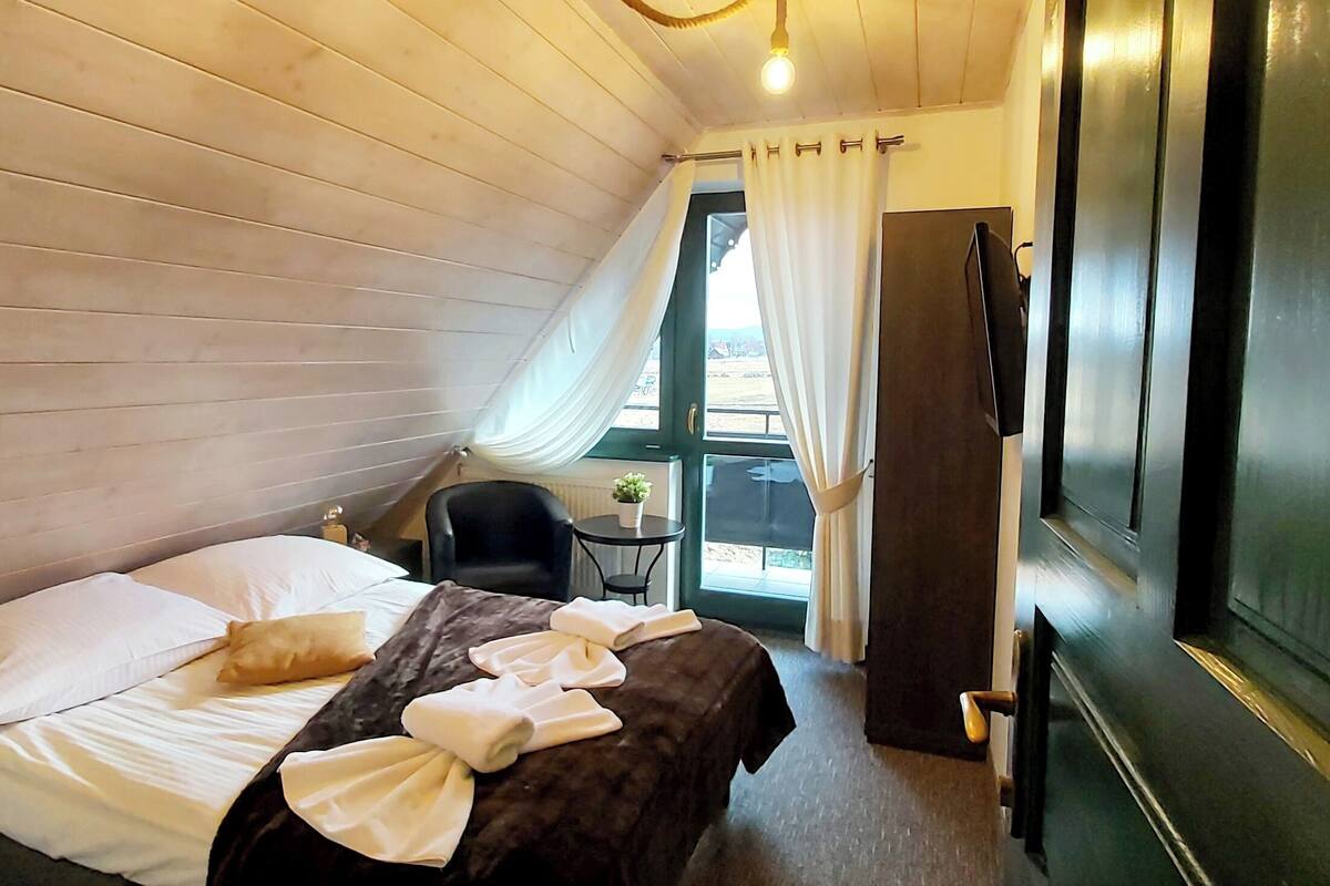 A room with a bed, a chair, a small table with a plant, and a TV on the wall.