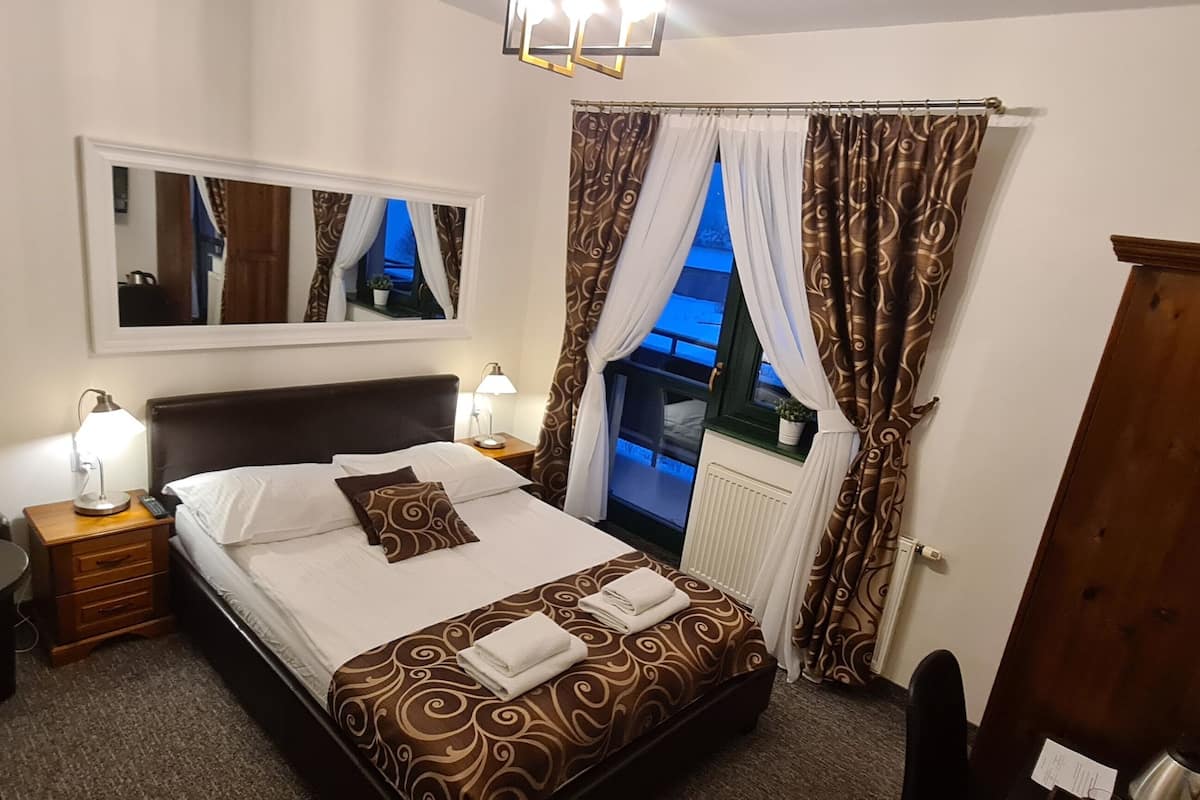 A hotel room with a bed, bedside lamps, a chair, a small table, a mirror, and a view of the outside through curtains.