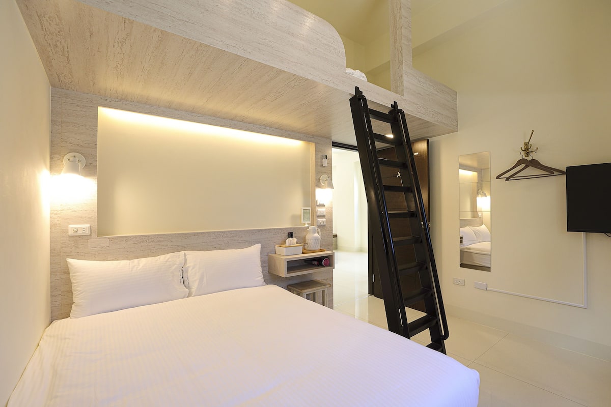 Quarto de hotel com cama, mesa de cabeceira, televisão fixada na parede e escada de madeira.