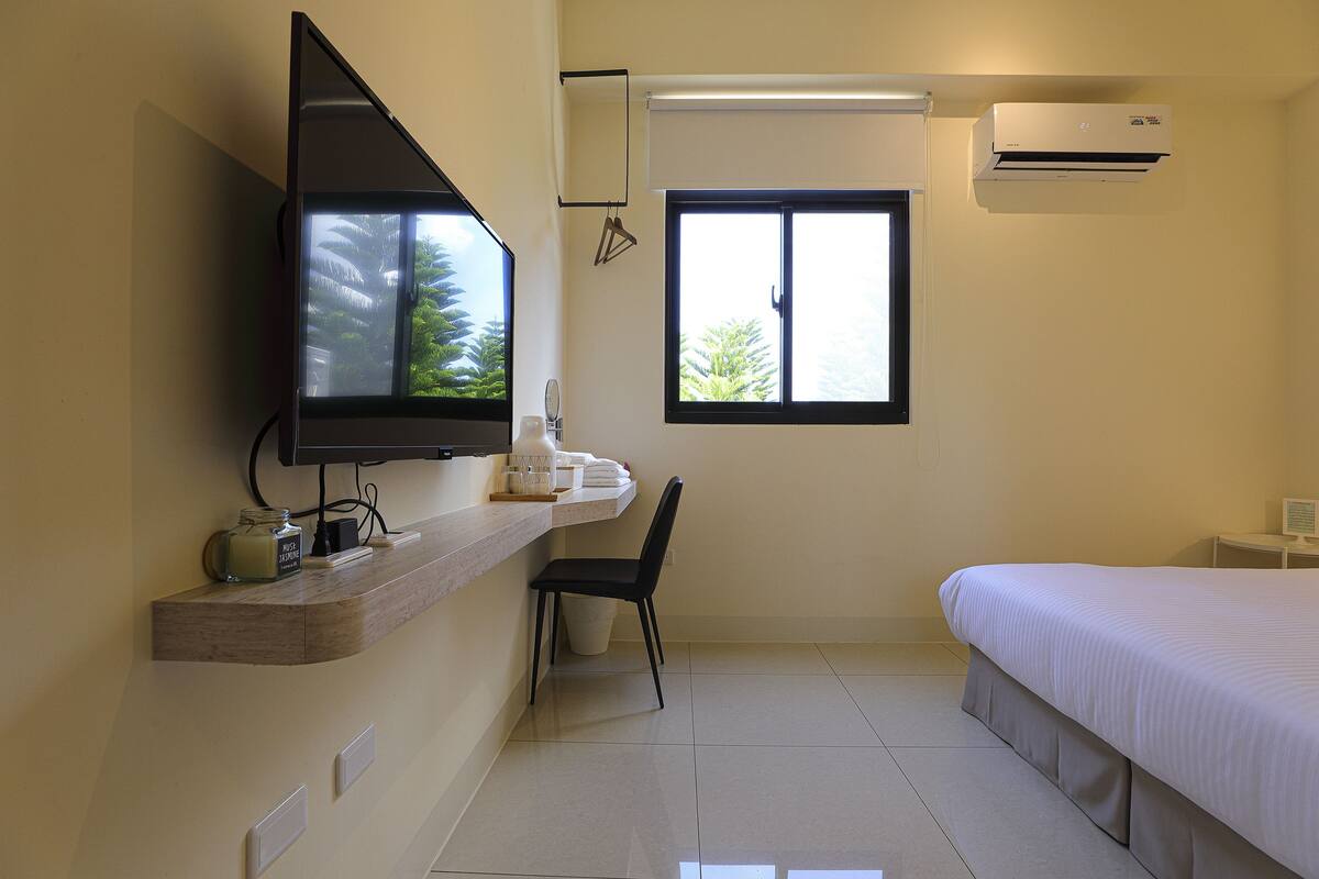 Quarto de hotel com cama, televisão fixada na parede, janela com vista para árvores e ar condicionado.
