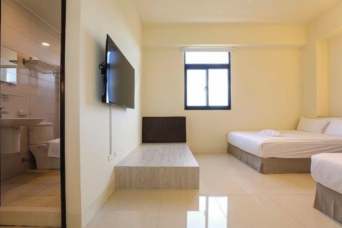 A modern hotel room with a flat-screen TV mounted on the wall, a small window, and a bed with a white comforter.