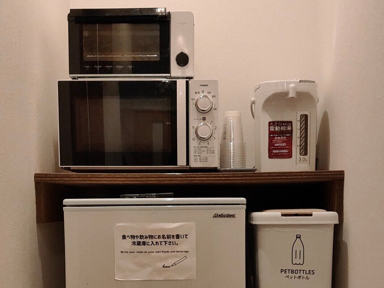A kitchenette with a microwave, toaster, and a small refrigerator. There is also a trash bin labeled 'PETBOTTLES' and a water dispenser.