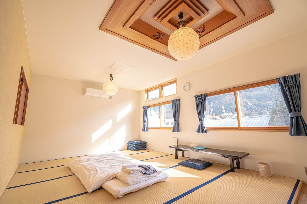 A room with tatami flooring, a bed with neatly folded blankets, a small table, a bench, and a window with curtains.