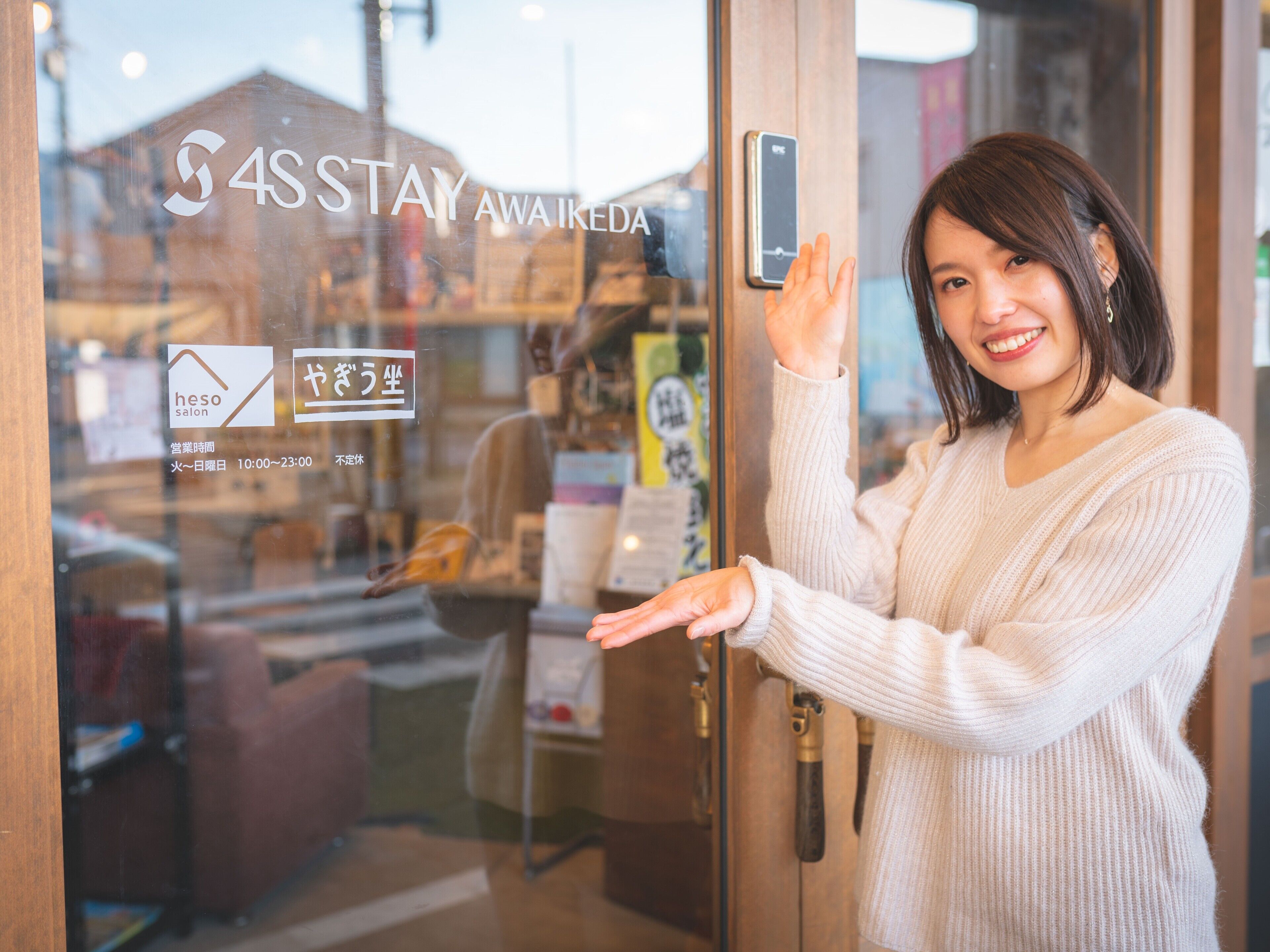 Una persona de pie frente a la entrada de un hotel, con un vidrio y un cartel que dice '4S STAY AWAKEDA', y se puede ver la distribución de las habitaciones con muebles en el interior.