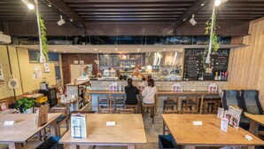 Interior de restaurante con mesas y sillas de madera, un mostrador con menú y plantas colgantes.