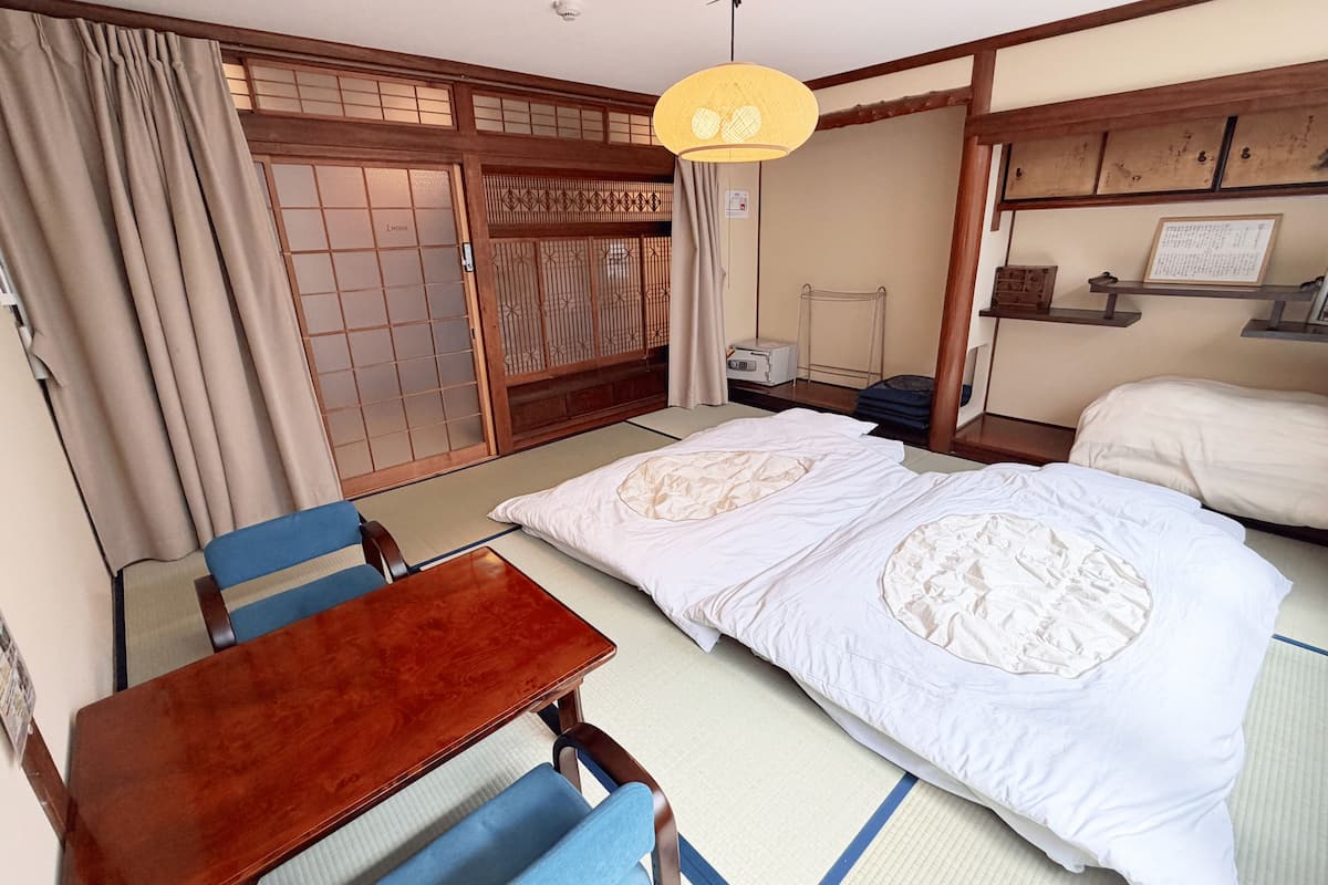 A traditional Japanese room with tatami flooring, a wooden table with blue chairs, two beds with white bedding, and a sliding door with a decorative screen.