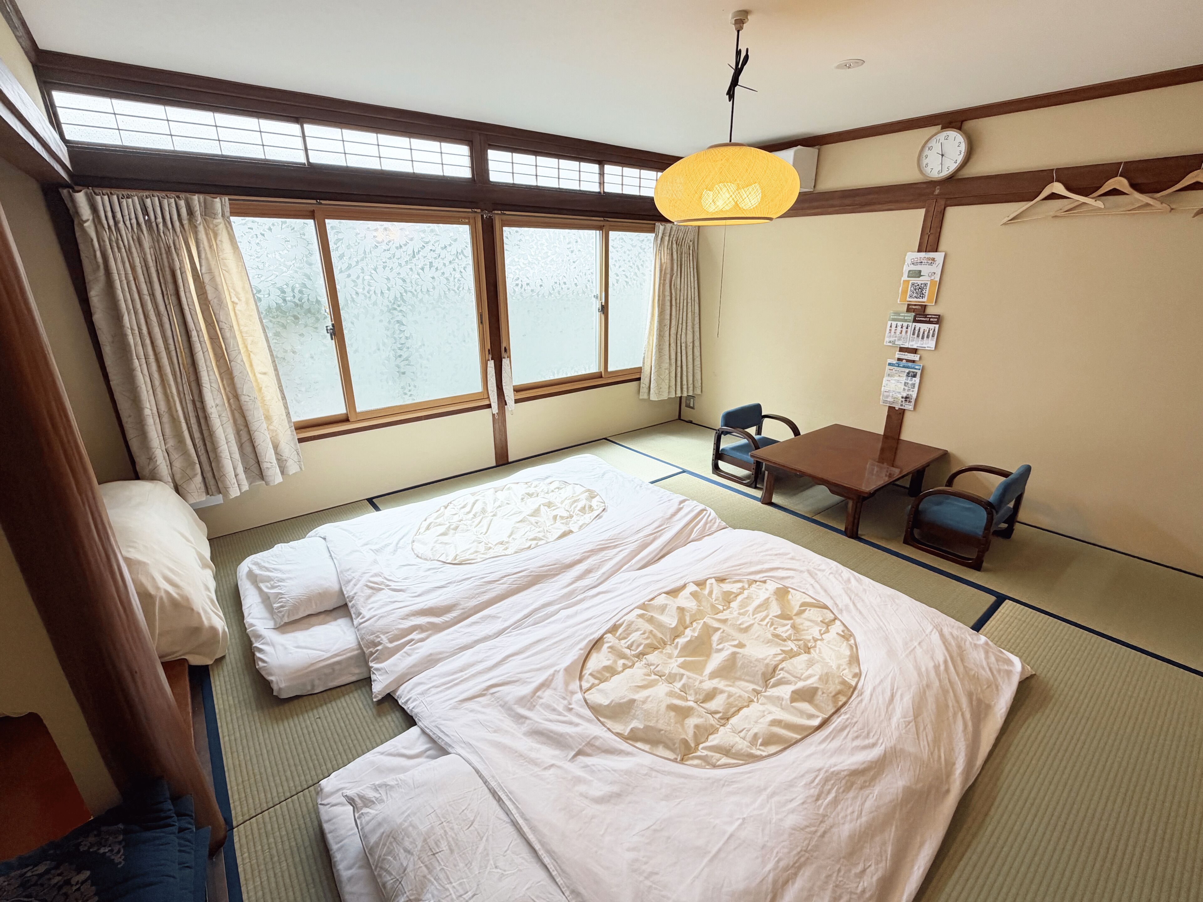 Una habitación japonesa tradicional con suelo de tatami, dos futones de tatami, una pequeña mesa con sillas, una lámpara colgante y un reloj en la pared.