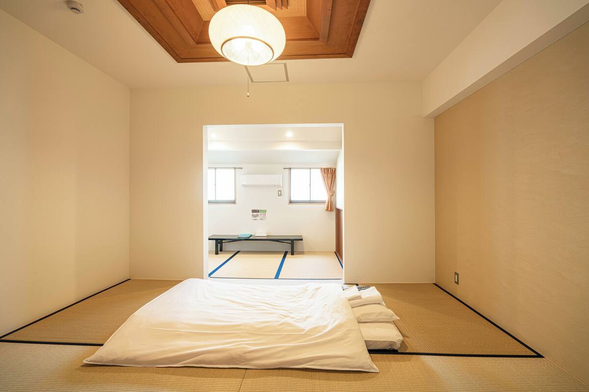 A tatami mat room with a bed, a bench, and a view into another room.