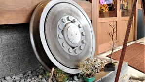 Una rueda de metal, posiblemente parte de un vehículo, apoyada contra un edificio con una pequeña planta en maceta cerca.