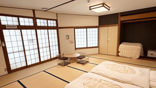 Una habitación japonesa tradicional con suelo de tatami, una mesa baja de tatami, dos camas de tatami, puertas correderas con paneles de vidrio y un armario.