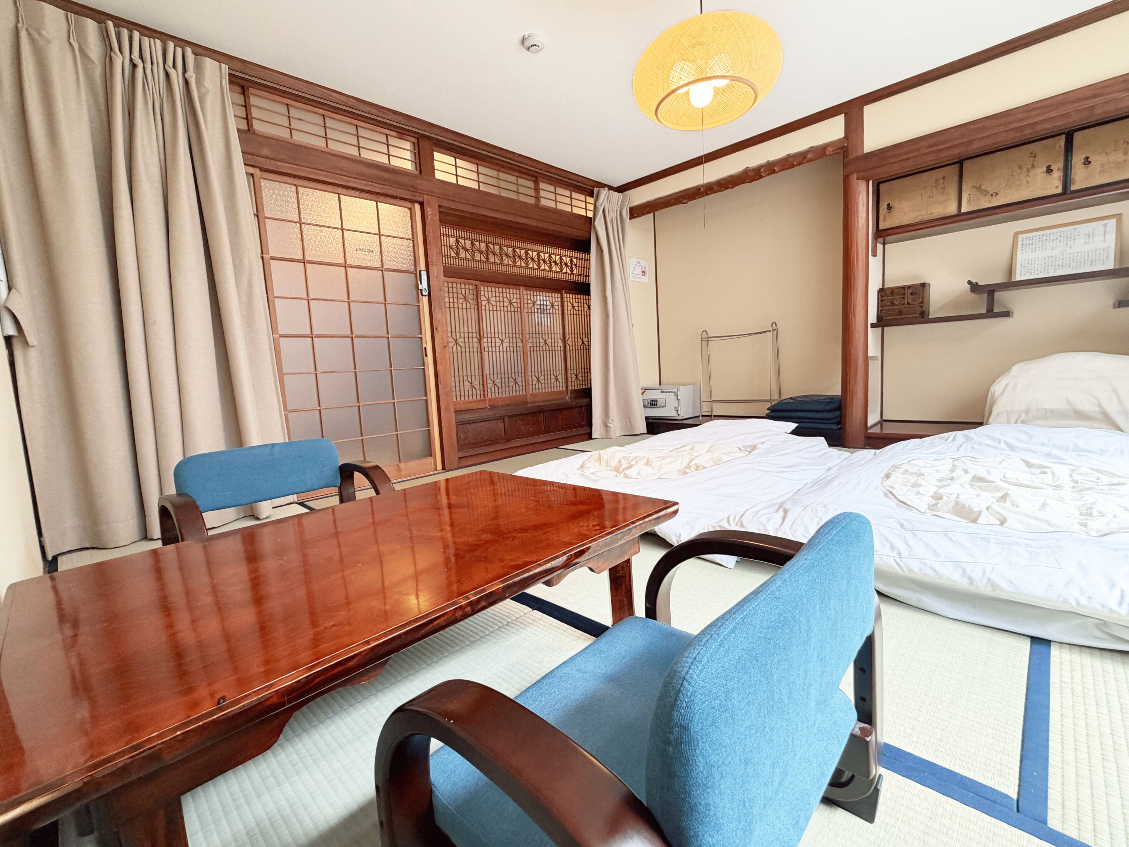 A traditional Japanese room with tatami flooring, a wooden table, a bed with white bedding, and a blue chair.