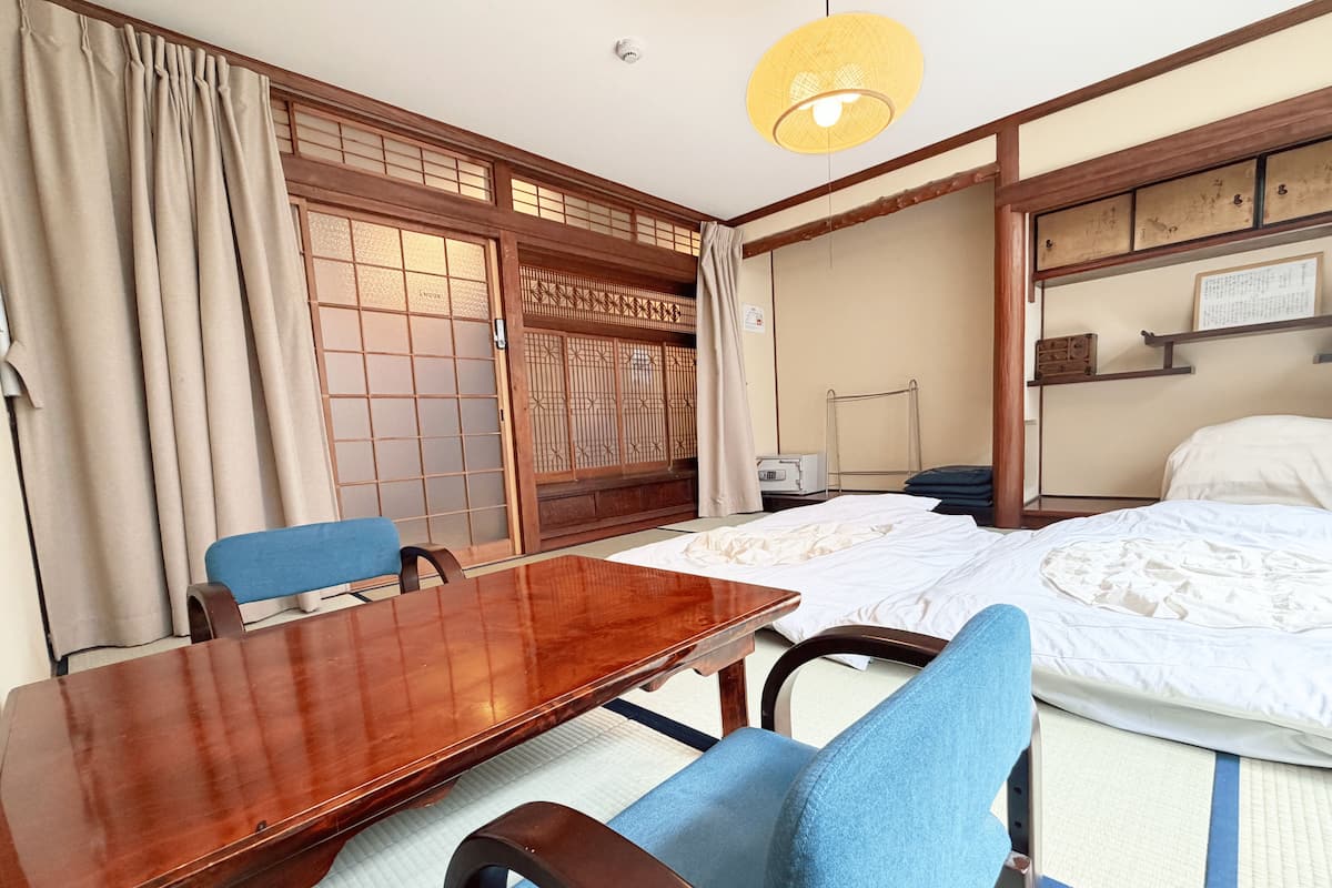 A traditional Japanese room with tatami flooring, a wooden table, a bed with white bedding, and a blue chair.