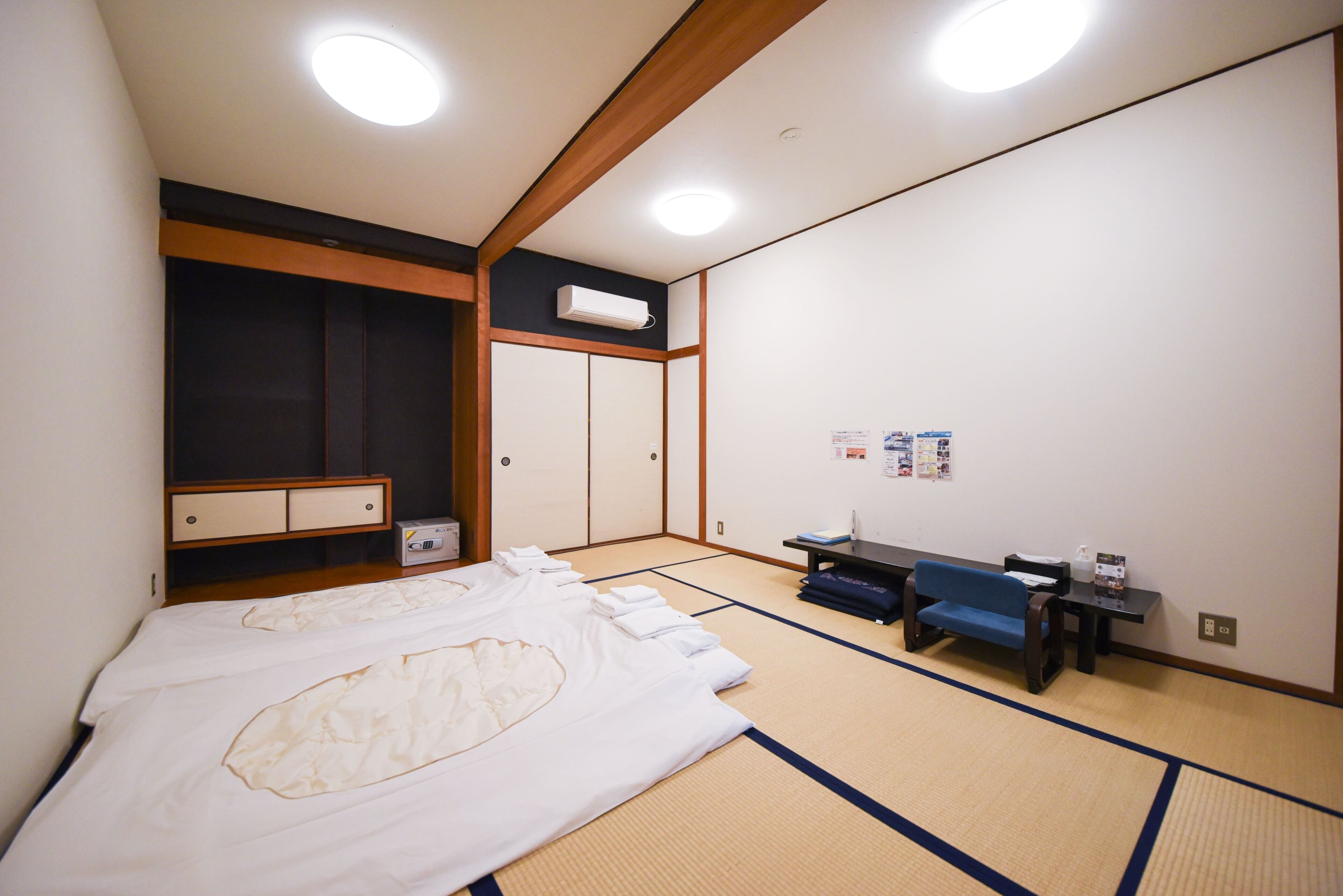 A traditional Japanese room with tatami flooring, a bed with white bedding, a blue sofa, a small table, and a wall-mounted air conditioner.