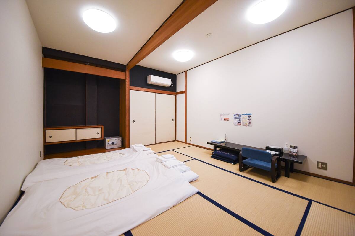 A traditional Japanese room with tatami flooring, a bed with white bedding, a blue sofa, a small table, and a wall-mounted air conditioner.