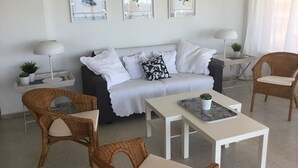 Un salon moderne avec un canapé blanc, deux fauteuils en osier, une table basse blanche et des œuvres d’art encadrées accrochées au mur.