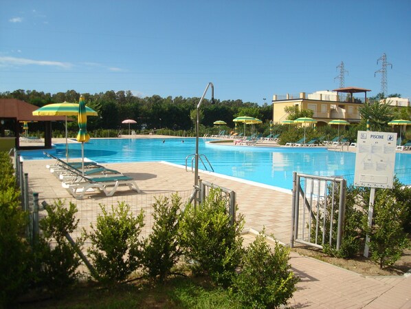 Una piscina con tumbonas y sombrillas, rodeada de vegetación y una zona vallada.