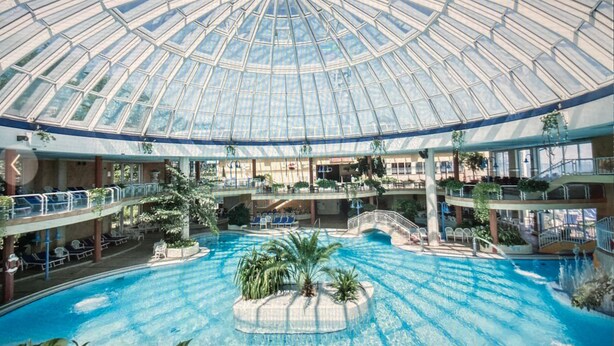 A large indoor swimming pool with a glass dome ceiling, multiple levels with balconies, and various seating areas. - App. with elevator + gr. Balcony + WLAN + 2 bicycles + beach chair package possible (Travemünde)