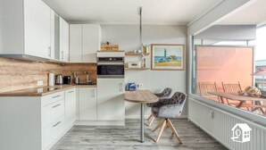 Una cucina moderna con mobili bianchi, un piano di lavoro in legno e un'area pranzo con tavolo e sedie. Appeso al muro c'è un quadro e, attraverso un'ampia finestra, si intravede l'esterno.