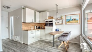A compact kitchen with white cabinets, a wooden countertop, and a built-in oven. There's a dining area with a table and a chair, and a window with a view of a beach. - App. with elevator + gr. Balcony + WLAN + 2 bicycles + beach chair package possible (Travemünde)