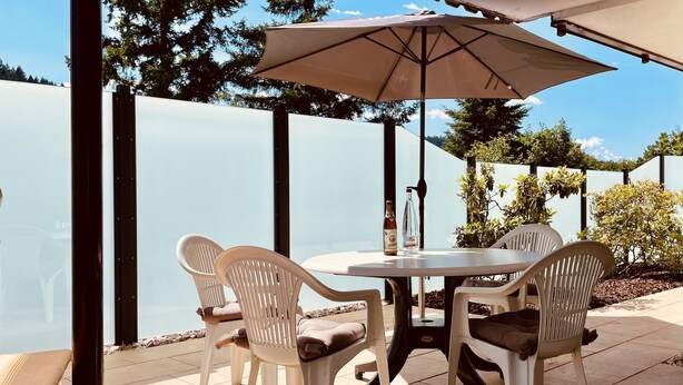 En udendørs terrasse med et bord, stole og en stor parasol, med udsigt over havet.