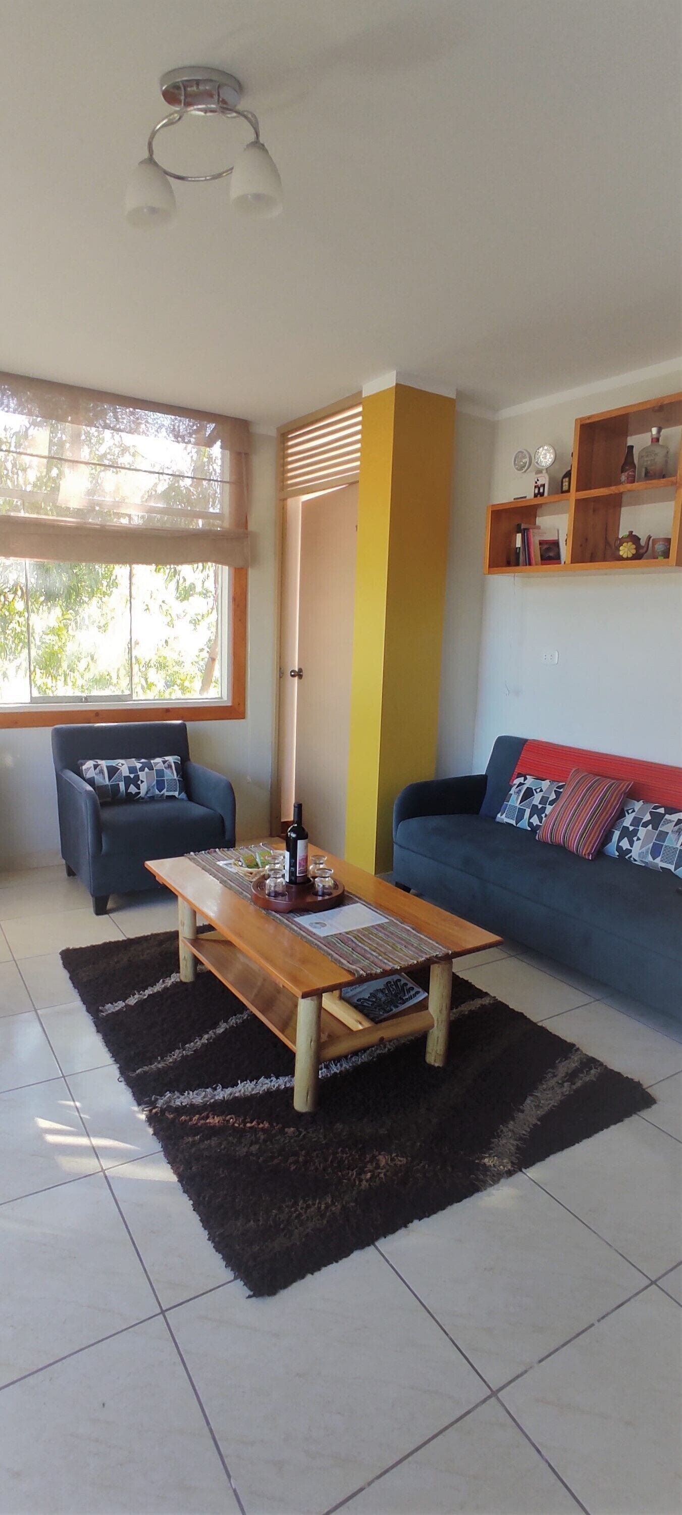 Un salon comprenant un canapé, une table basse, un fauteuil, une fenêtre avec des stores et une étagère ornée d’objets décoratifs.