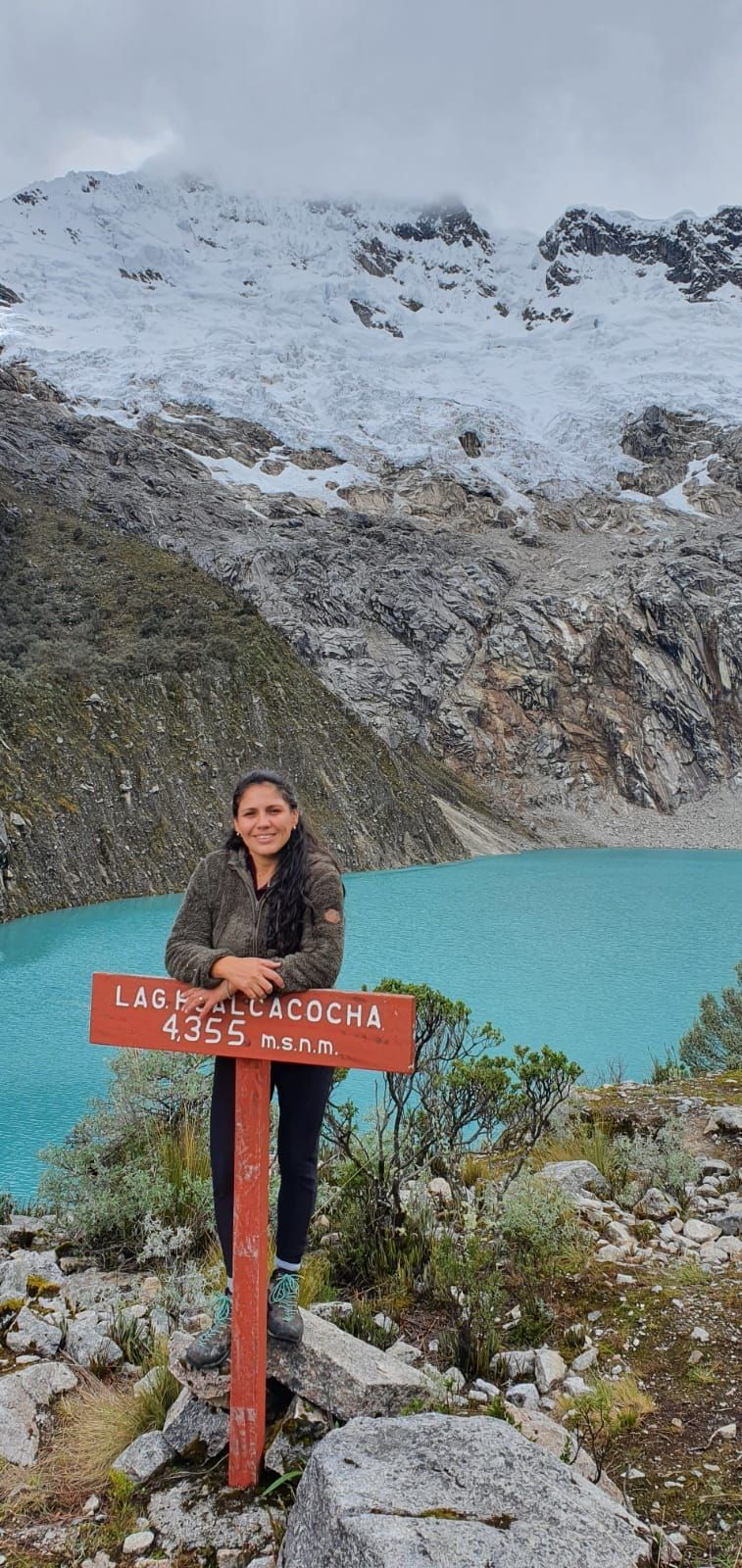 Una persona de pie junto a un poste de señalización, con una montaña y un lago al fondo.