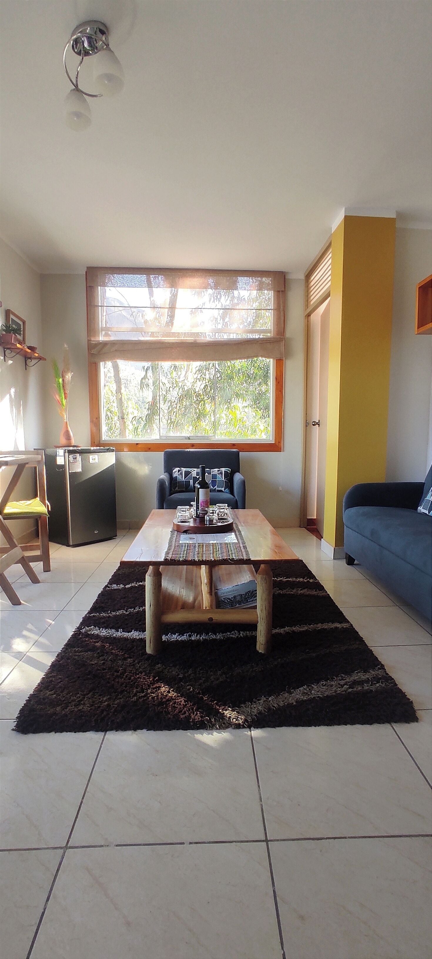 Un salón con una mesa de centro de madera, un sofá azul, un ventanal con vista a árboles y una pared amarilla.