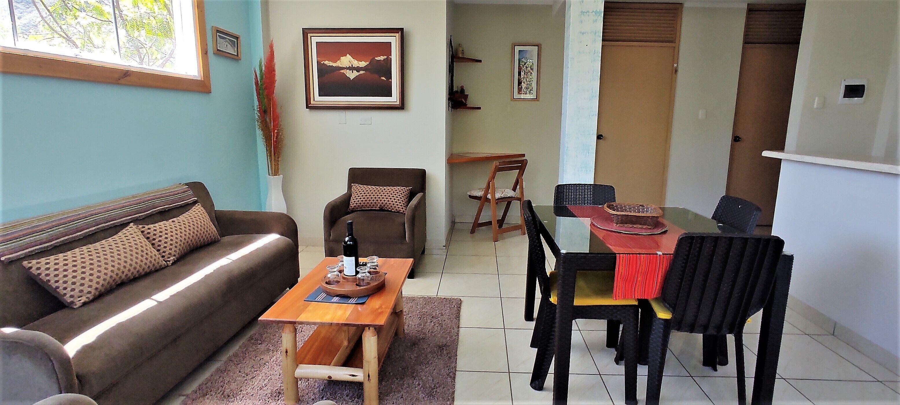 Uma sala de estar com um sofá, uma mesa de centro de madeira e uma mesa de jantar com cadeiras. Há uma pintura na parede e uma janela com vista para árvores.