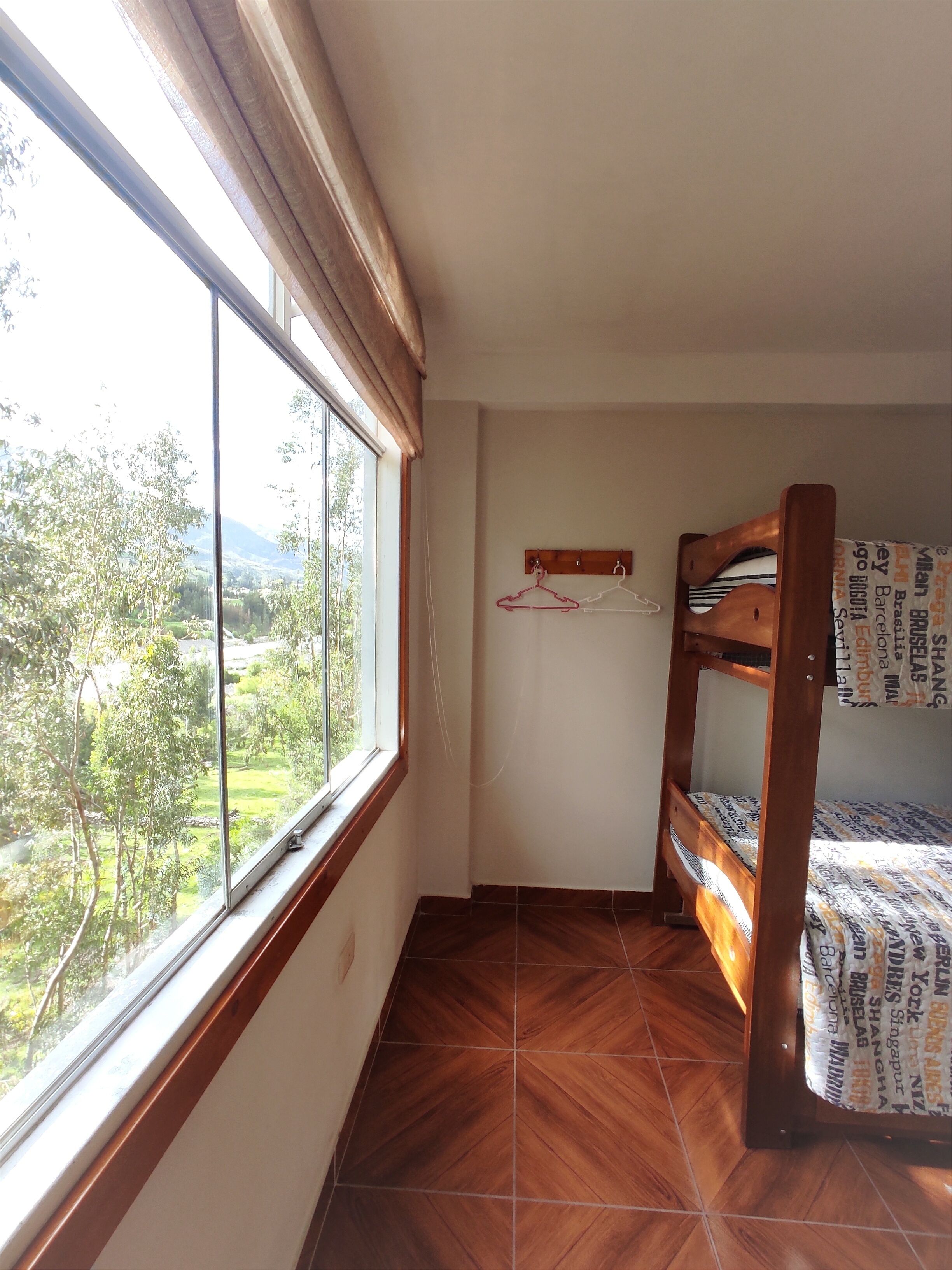 Una habitación con una cama litera de madera, una ventana con vistas a follaje, y un perchero en la pared.