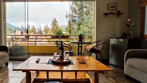 Una sala de estar con una mesa de madera, dos sofás, una mesita redonda y vistas a un balcón con plantas y una montaña al fondo.