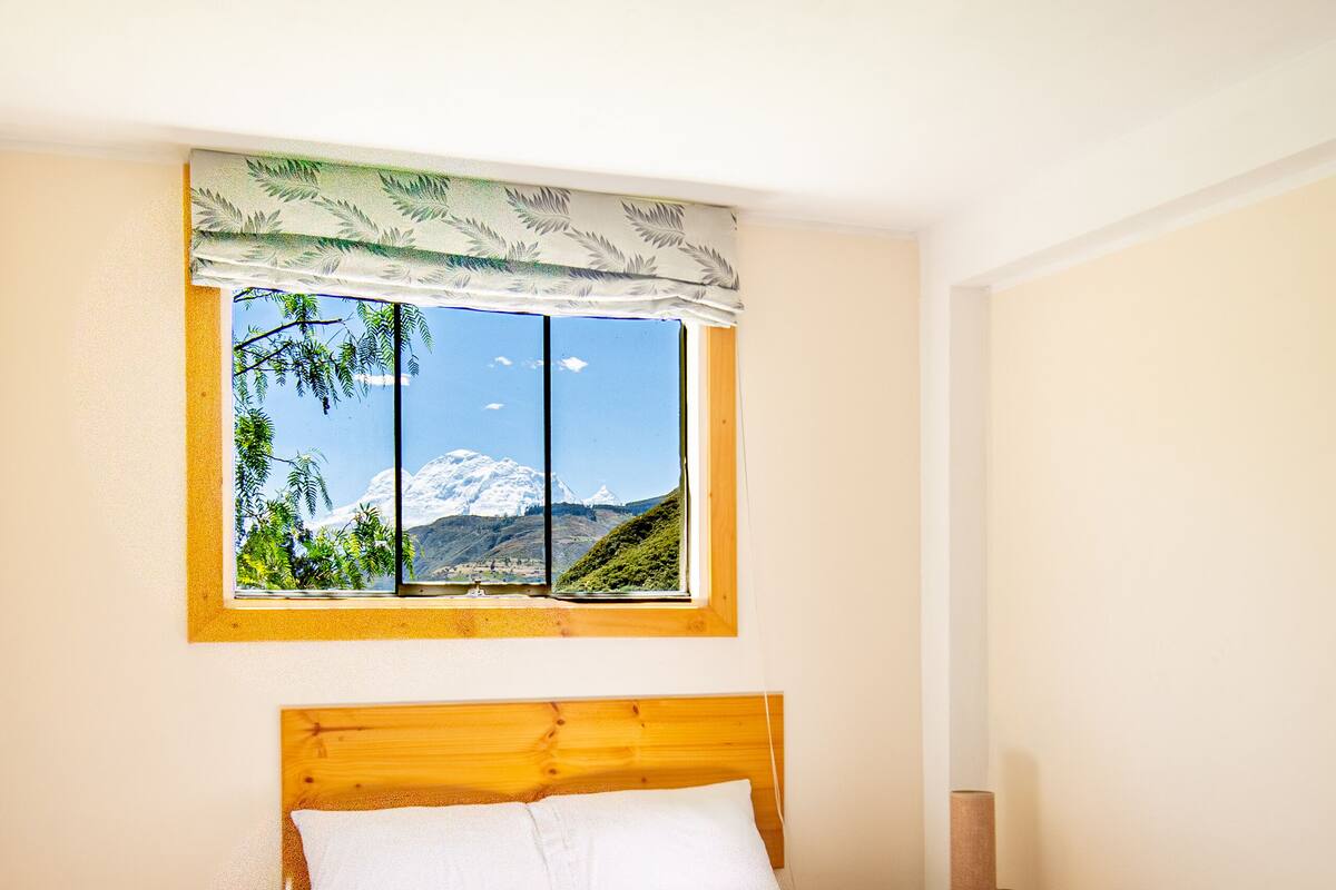 A bedroom with a wooden bed, a window with a view of a mountain, and a small bedside table with a lamp.