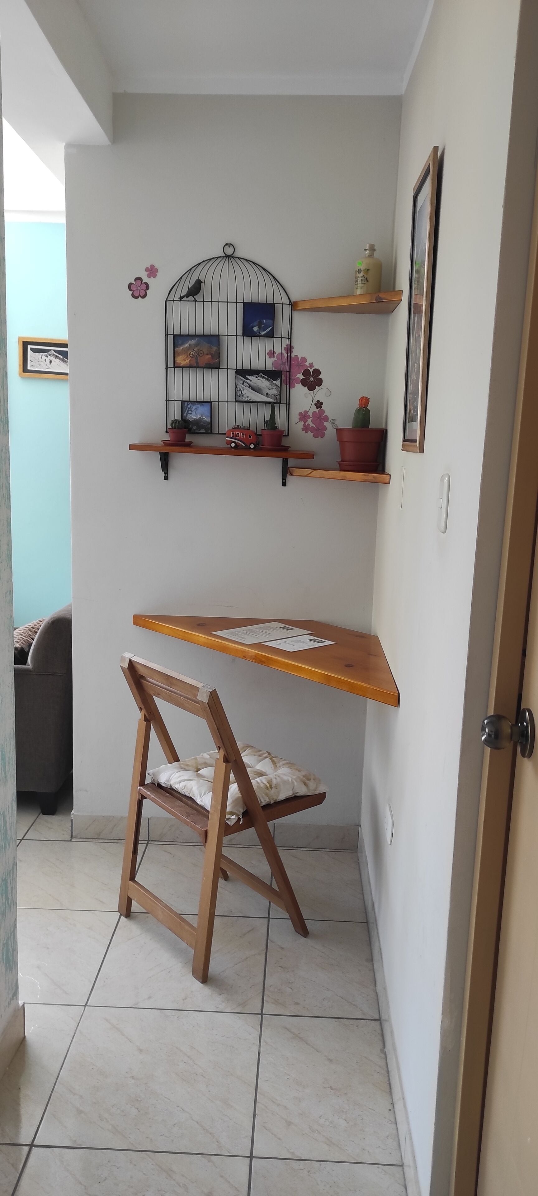 A corner room with a wooden desk, a chair, a wall-mounted shelf with potted plants, and a birdcage decoration.