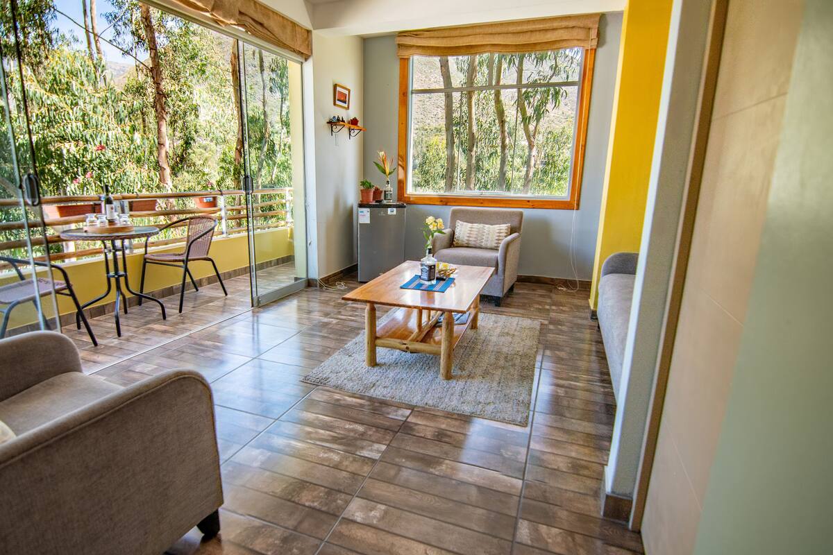 A spacious living area with a wooden coffee table, a sofa, and a dining table with chairs. A large window offers a view of trees and a balcony.