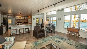 A spacious living area with a large window overlooking a body of water, a kitchen with wooden cabinets, and a dining area with a table and chairs. - The Sanctuary on Golden Lake (Pleasant Lake)