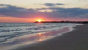 Ein Strand mit einem Sonnenuntergang über dem Ozean, sanfte Wellen umspülen das Ufer, und eine entfernte Landmasse zeichnet sich vor dem Himmel ab.