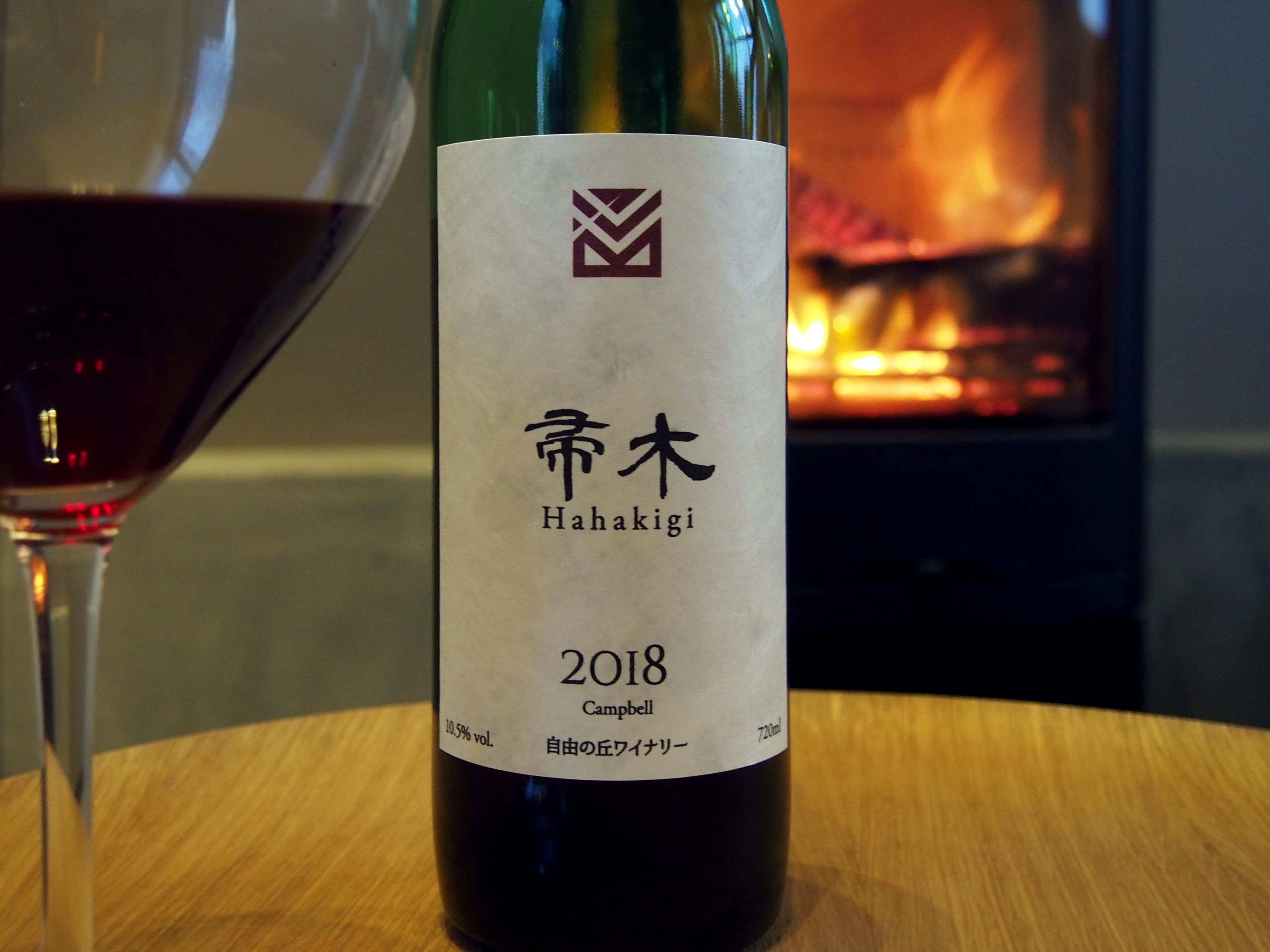 A bottle of wine with a label, placed on a wooden table with a glass of red wine beside it.