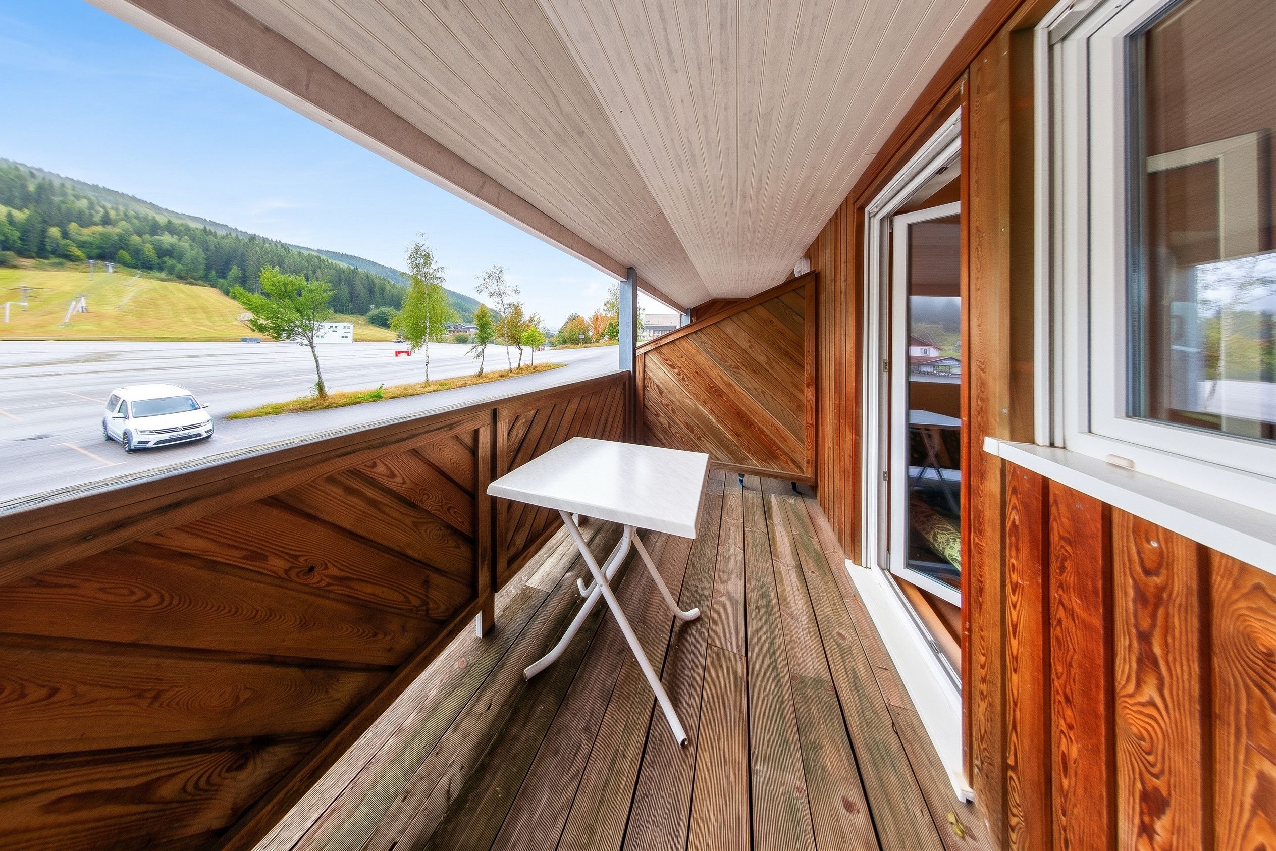 A wooden deck with a table and a window overlooking a road and trees.