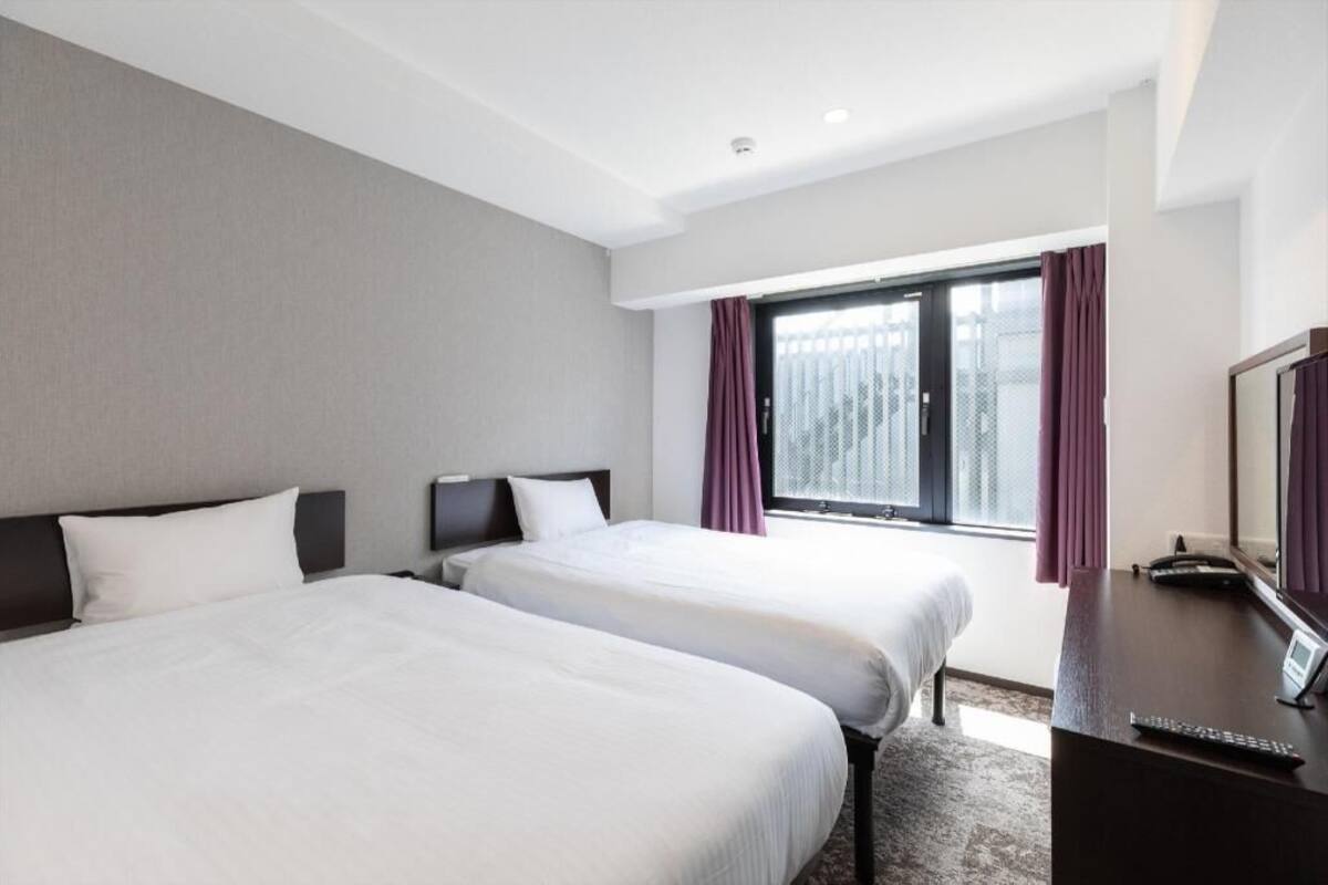 A hotel room with two beds, a desk, a TV, and a window with curtains.