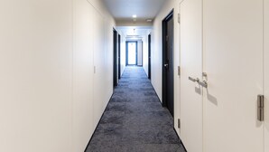 Un largo y estrecho pasillo con paredes blancas y una alfombra oscura. Hay puertas a ambos lados y una lámpara en el techo.