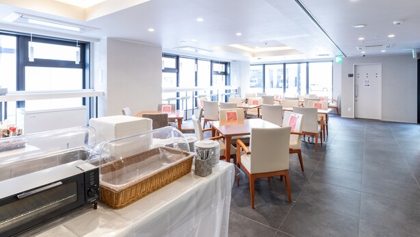 Un comedor amplio y luminoso con varias mesas y sillas, ventanas grandes y un sistema de buffet con un microondas y cestas.