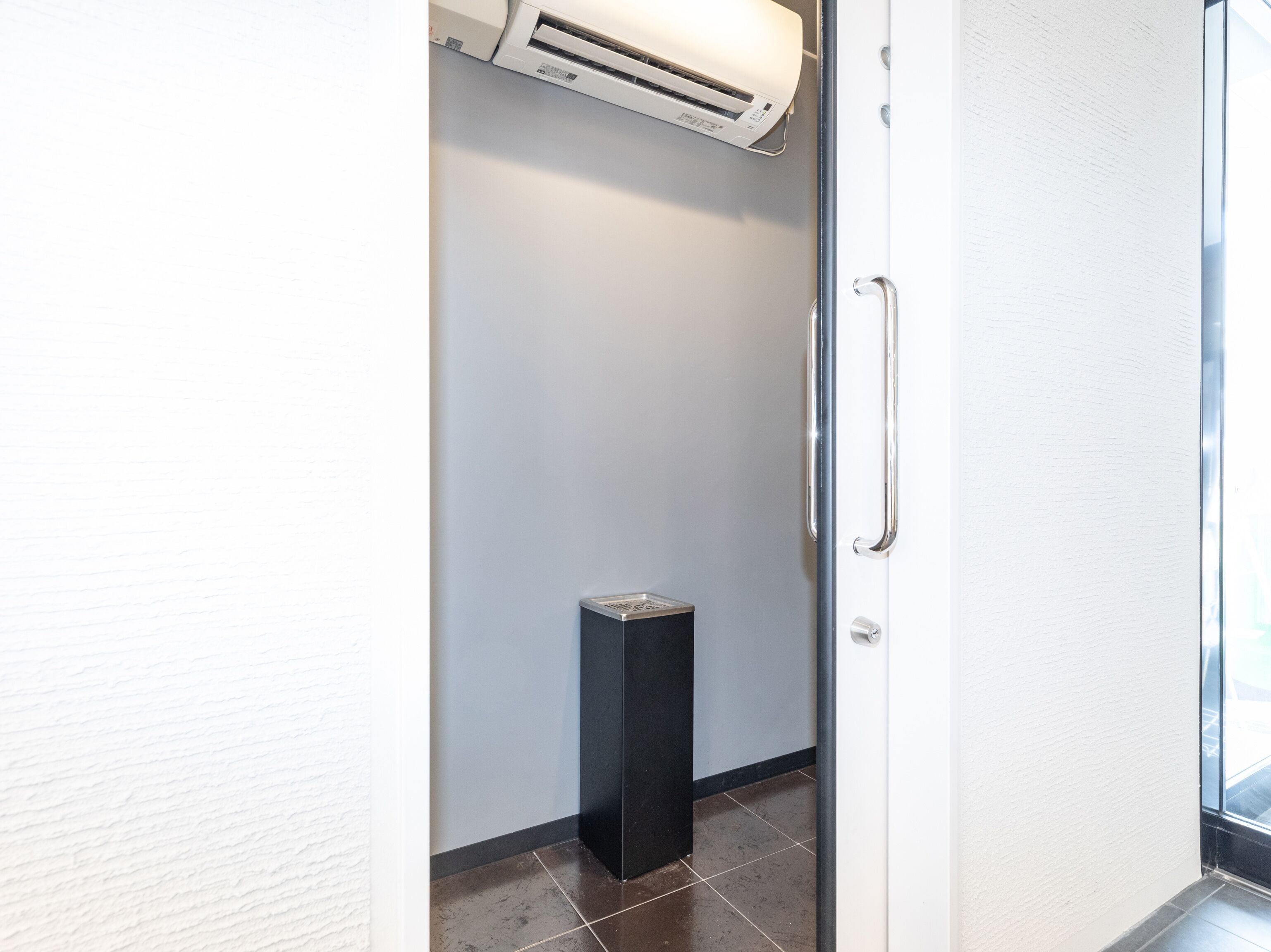 A narrow hallway with a door on the right, a trash bin, and an air conditioning unit mounted on the wall.