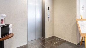 Un ascensor moderno con pantalla digital, un microondas sobre un pedestal y un cartel en un pasillo con paredes texturizadas.